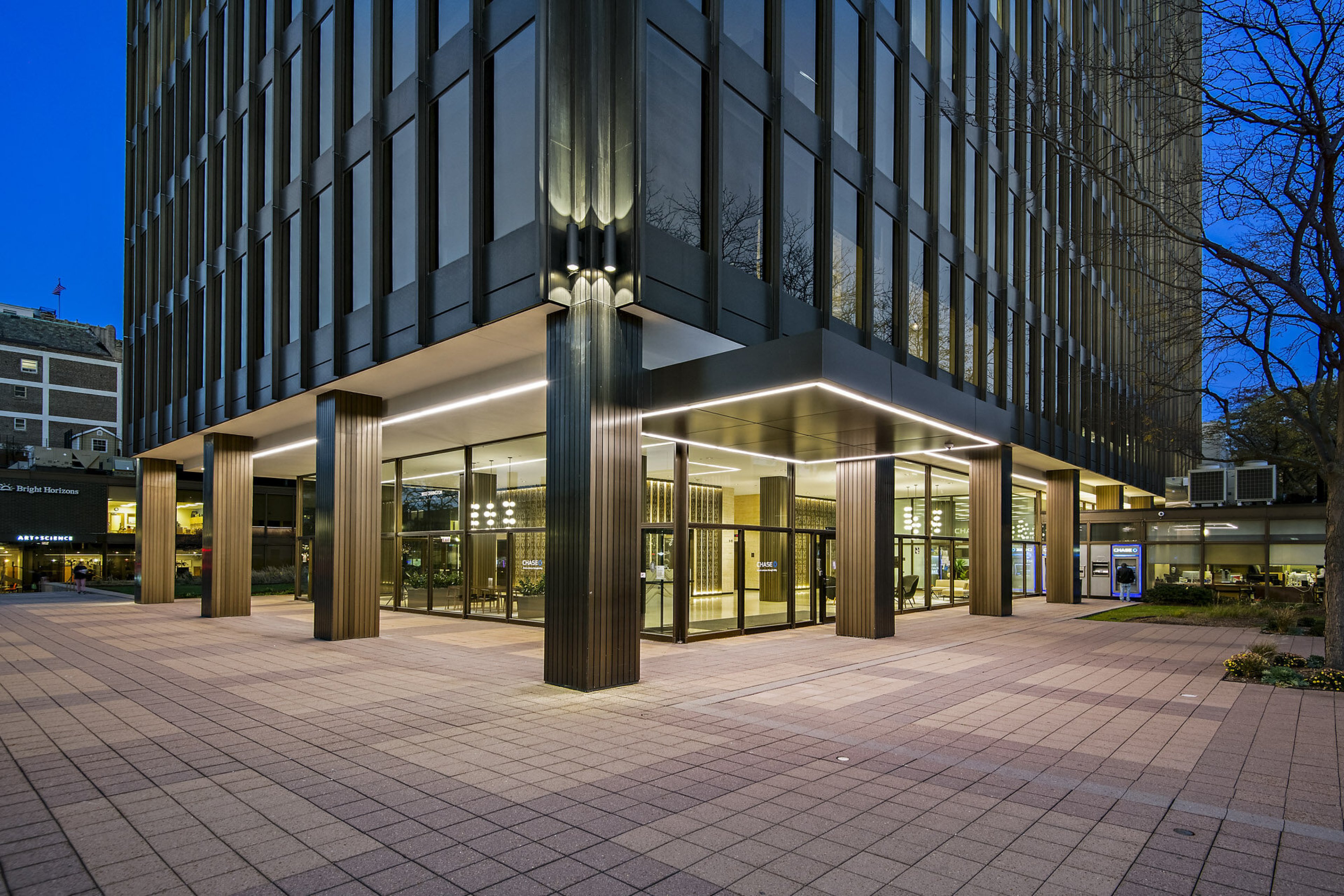 Illuminated glass lobby entrance of Orrington Plaza at dusk, framed by fluted bronze columns and a paved plaza